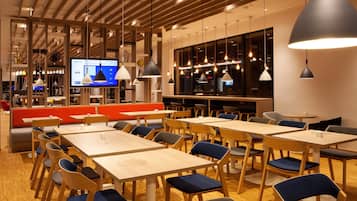 Un restaurante moderno con mesas y sillas de madera, una pantalla de televisión grande e iluminación colgante.