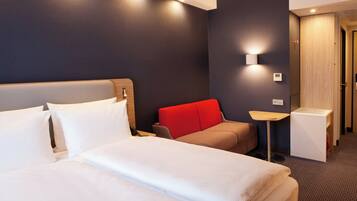A hotel room with a bed, a red chair, a small table, and a wall-mounted light.