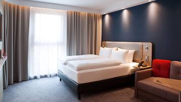 A modern hotel room with a large bed, a bedside table with a lamp, a red chair, and a small wooden table.