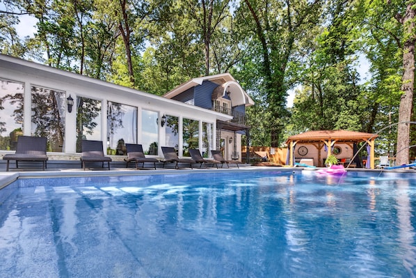 Une piscine avec des chaises longues, un espace de détente couvert et un bâtiment doté de grandes fenêtres et d’un balcon.