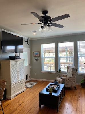 A living room with a ceiling fan, a flat-screen TV, a white cabinet, a patterned armchair, and a wooden coffee table.