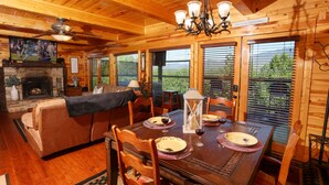 A wooden cabin interior with a dining table set for four, a fireplace, and a cozy living area with a sofa and a television.