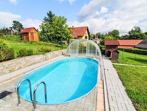 Une piscine dotée d’une enceinte en verre, entourée d’une terrasse en briques et d’une pelouse agrémentée d’arbres, avec des maisons en arrière-plan.