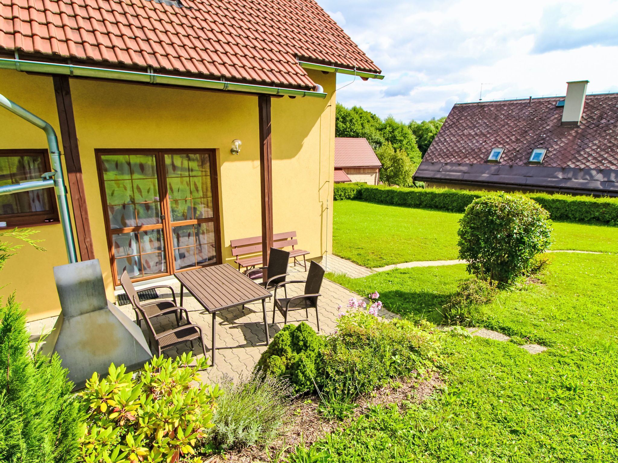 Un bâtiment jaune avec un toit recouvert de tuiles, une terrasse avec une table et des chaises, et une pelouse bien entretenue.