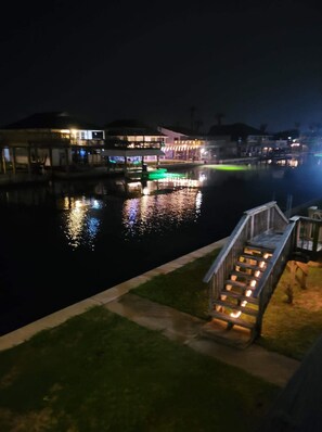 Une propriété située au bord de l’eau, de nuit, avec des bâtiments illuminés dont les reflets se mirent dans l’eau.