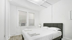 A bedroom with a bed, white walls, a window with blinds, and a lamp. - Modern Beachfront Suite 206 (Ocean City)
