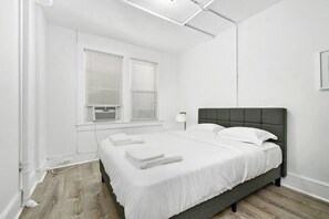 Une chambre à coucher avec un lit, des murs blancs, une fenêtre avec des stores et une lampe.