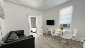 A modern room with a grey sofa, a dining table with chairs, a TV mounted on the wall, and a window with blinds. - Modern Beachfront Suite 206 (Ocean City)