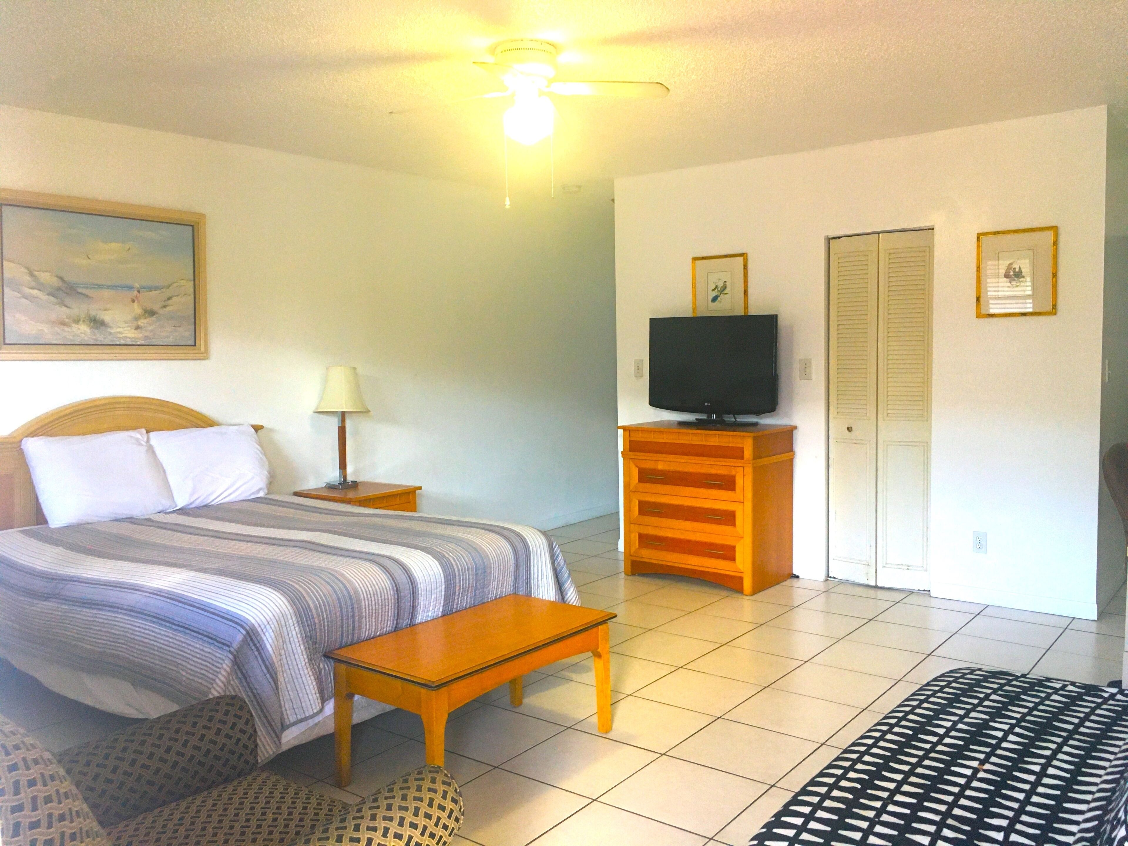 Una habitación de hotel con una cama, una mesita, un televisor sobre un soporte de madera, una lámpara y un ventilador de techo.