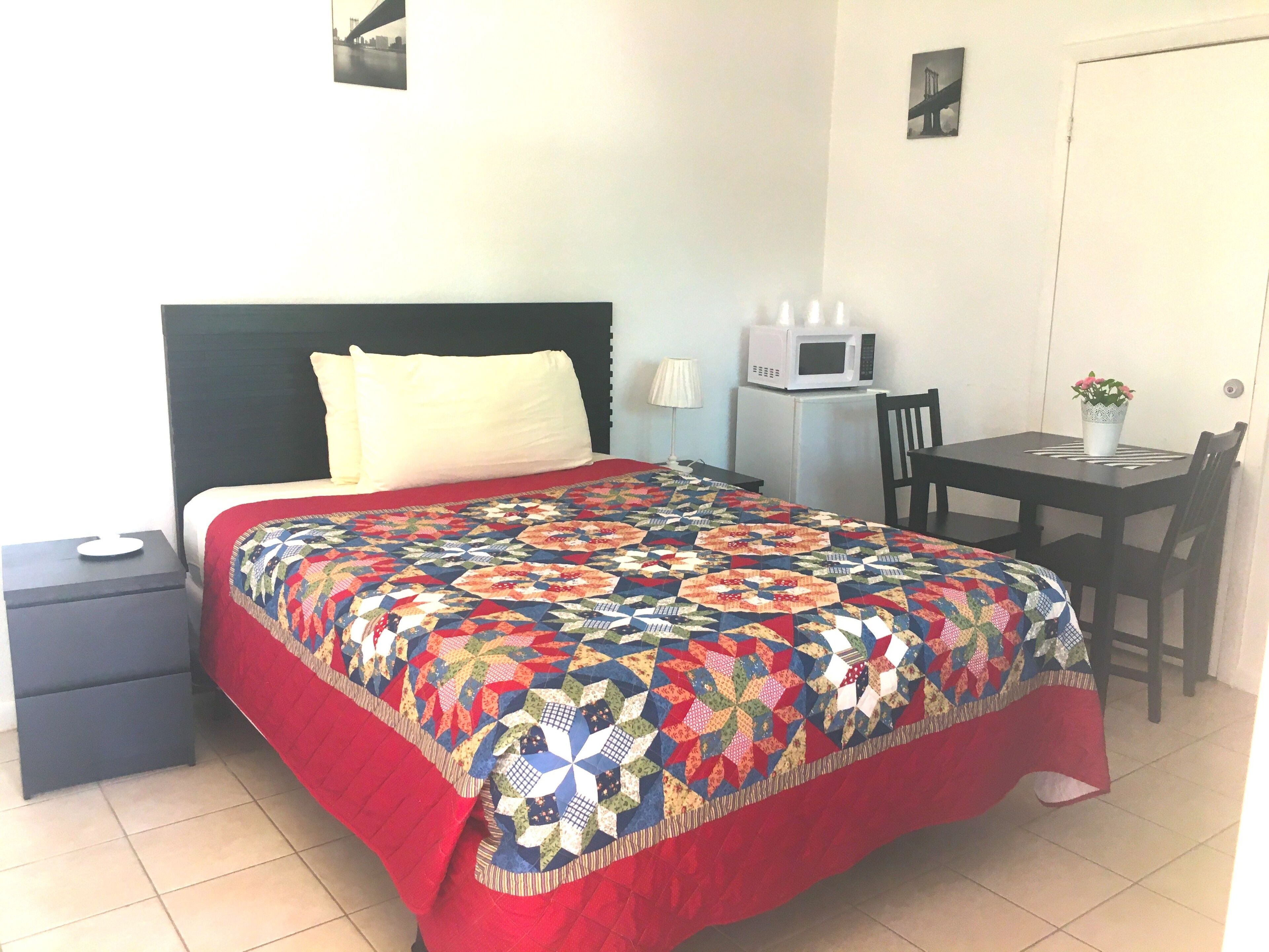 Una cama bien hecha con un colcha colorida, una mesita de noche pequeña, un escritorio con una silla, un microondas y una planta en maceta.