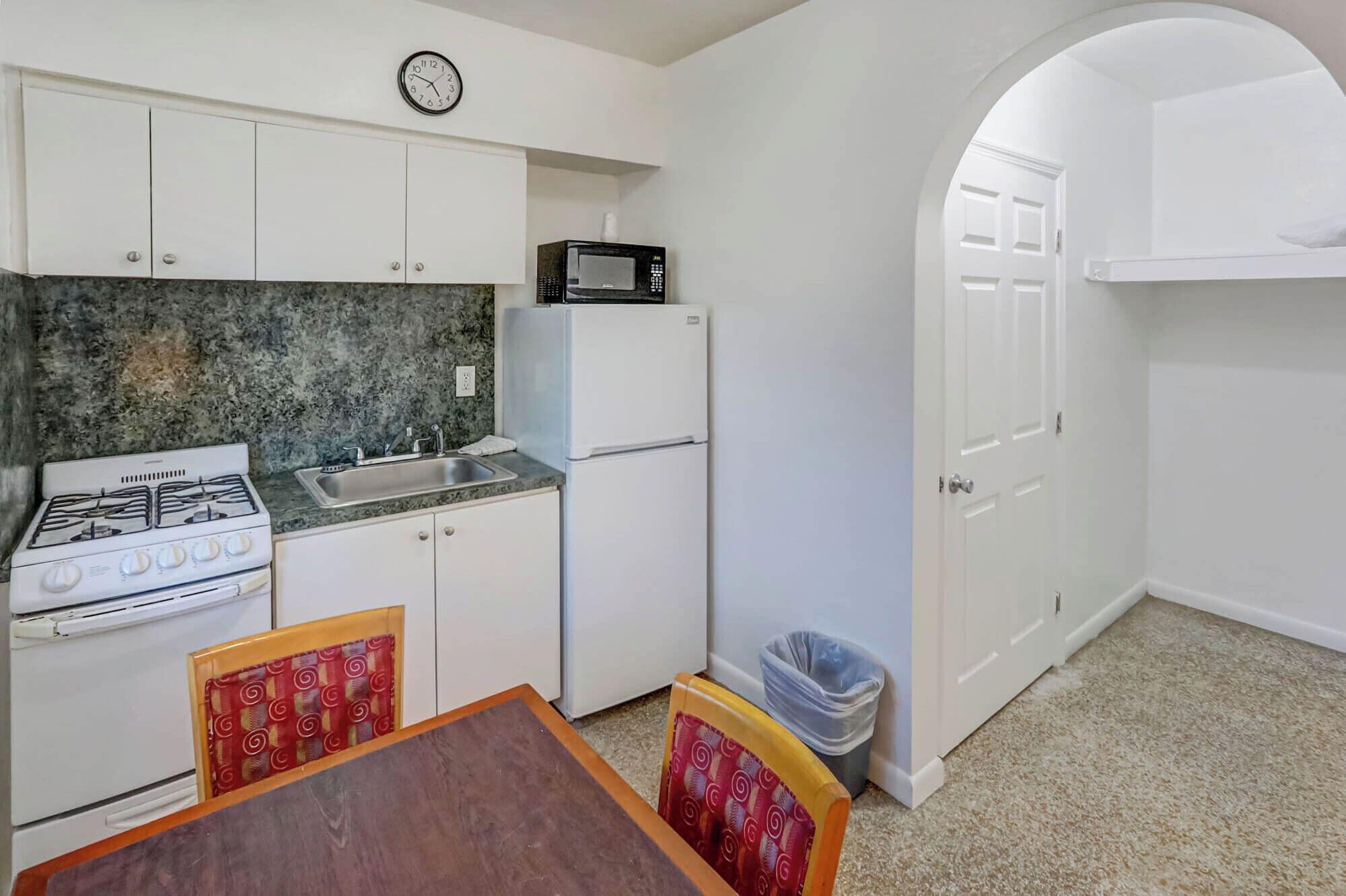 A compact kitchen with white cabinets, a gas stove, a microwave, and a sink. There is a dining table with chairs and a trash bin.