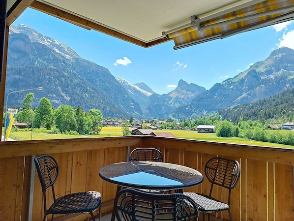 Un balcon avec une table et des chaises, donnant sur un paysage montagneux parsemé de maisons et de verdure.