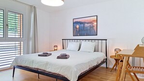 A bedroom with a bed, bedside lamps, a table, a chair, a window with blinds, and a wall-mounted picture.