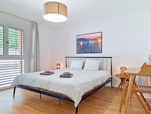 Une chambre à coucher comprenant un lit, des lampes de chevet, une table, une chaise, une fenêtre avec des stores et un tableau accroché au mur.