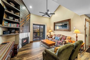 A cozy living room with a fireplace, a bookshelf, a sofa, and a view of the outdoors through a glass door.