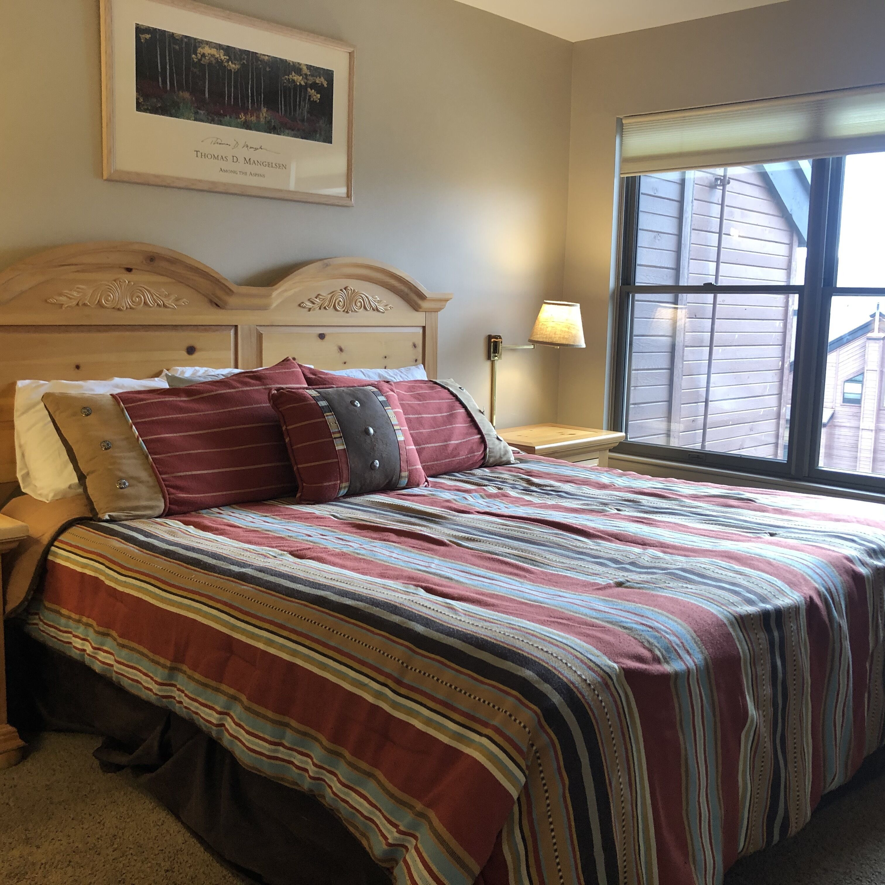 A bedroom with a wooden headboard, a large window with blinds, a bedside lamp, and a framed picture on the wall.