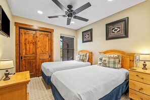 Habitación de hotel con dos camas, armario de madera, ventilador de techo, cuadros enmarcados, lámparas de noche y una mesita.