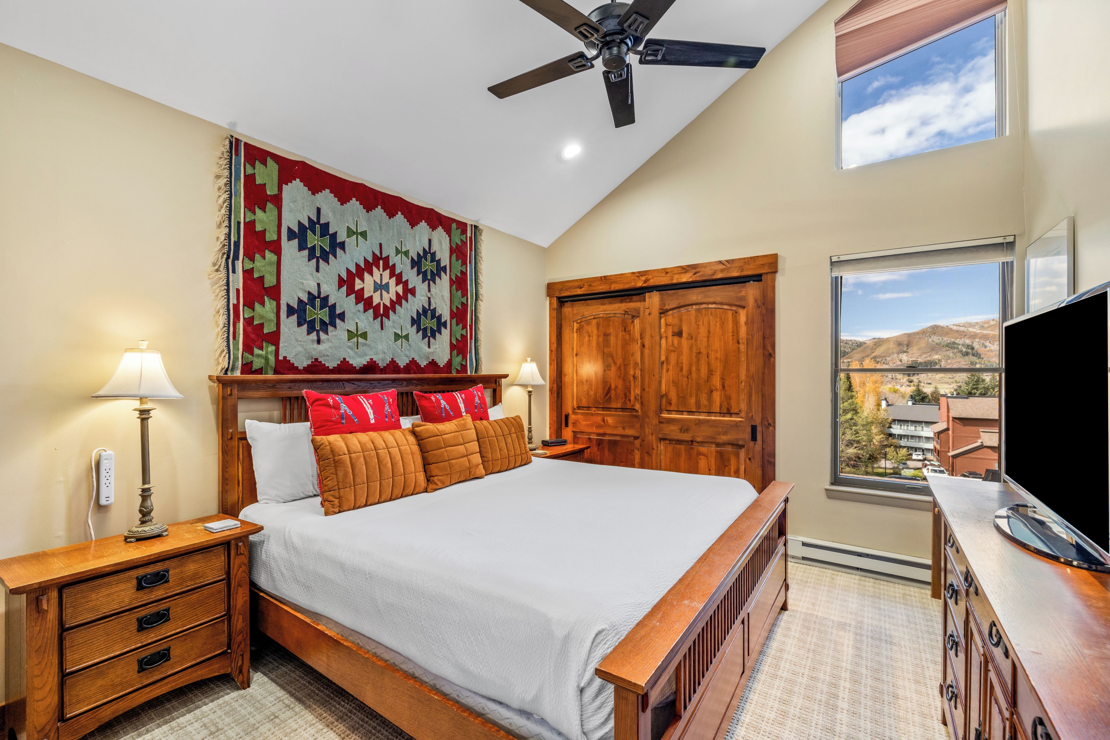 A bedroom with a bed, nightstands, a wooden wardrobe, a ceiling fan, and a window with a view.