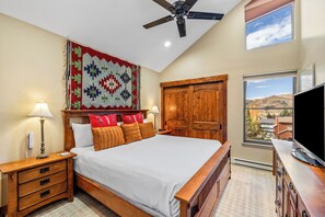 Une chambre à coucher comprenant un lit, des tables de chevet, une armoire en bois, un ventilateur de plafond et une fenêtre donnant sur un beau paysage.