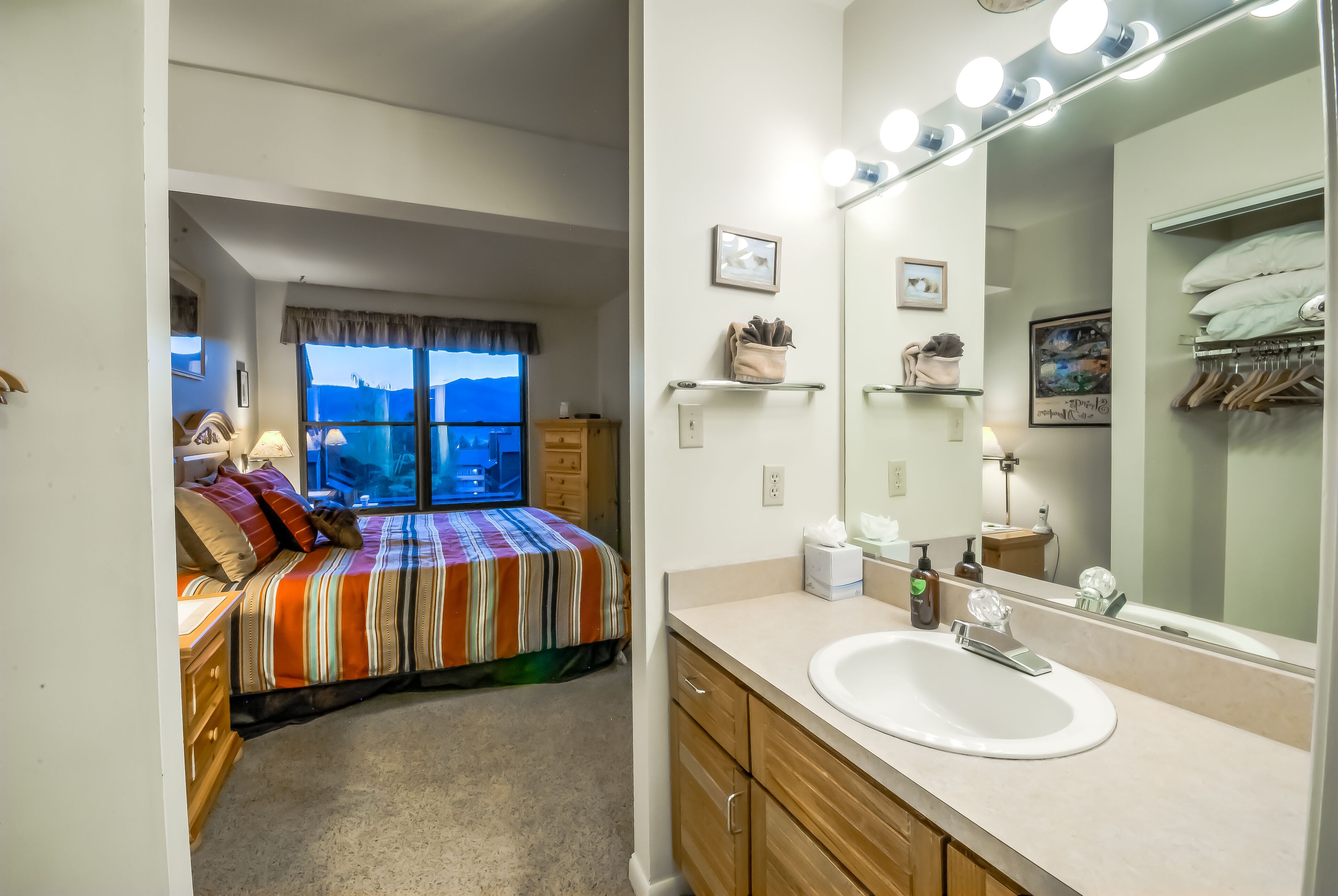 Une chambre avec un lit, une fenêtre donnant sur un beau paysage, une salle de bain avec un lavabo et un miroir, et un placard rempli de vêtements.