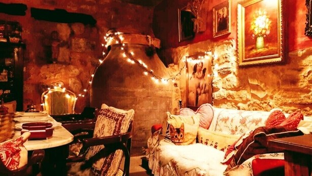 A cozy room with a rustic stone wall, a large ceramic pot, a wooden table with chairs, a sofa with patterned cushions, and a framed picture on the wall. - Romantic getaway to Valeria (Valeria)