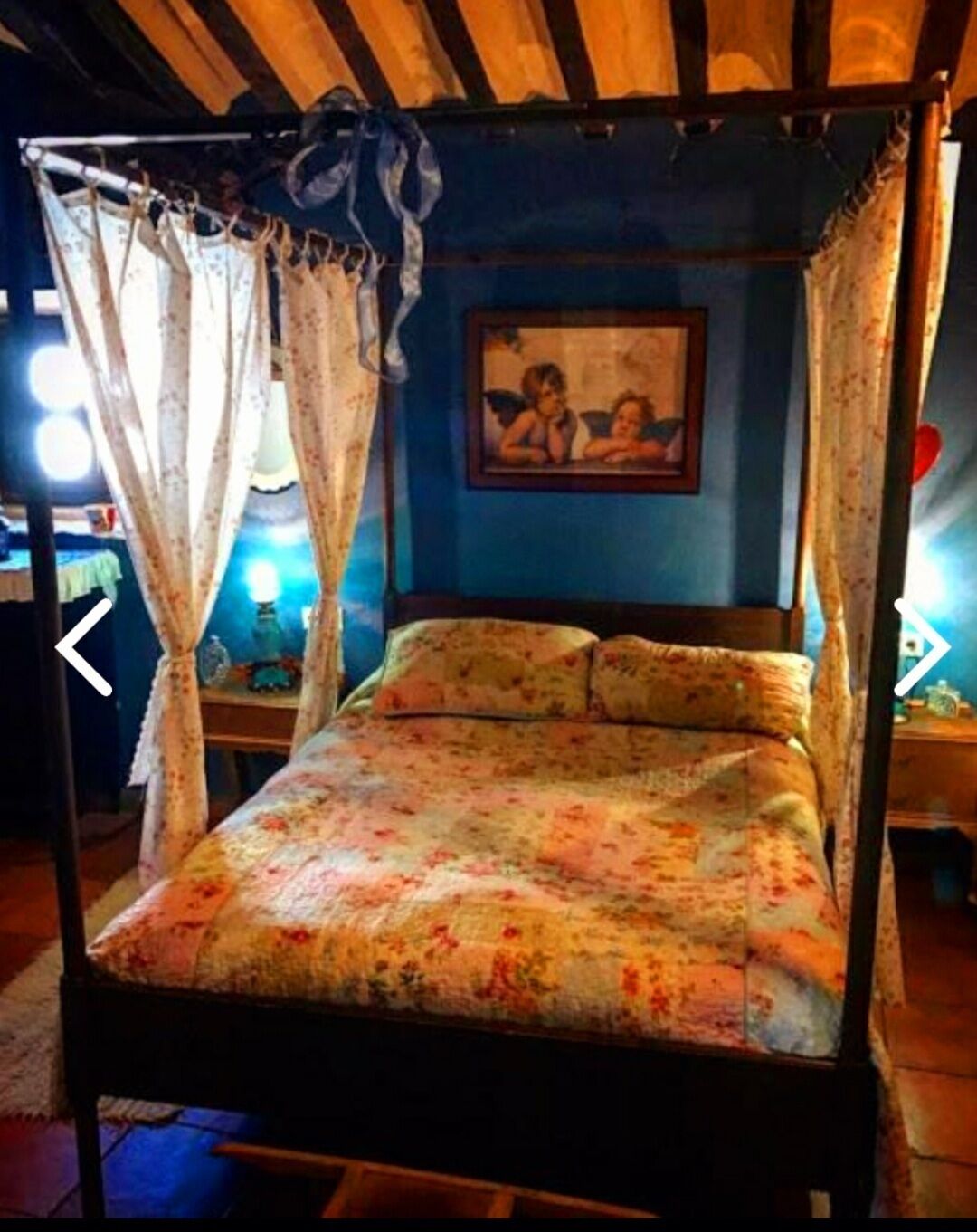 Una cama con dosel, ropa de cama floral, un cuadro colgado en la pared y una mesita de noche con lámpara.