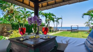 A wooden table with two red drinks and purple flowers, set against a backdrop of a clear blue sky and ocean view.