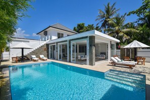 Et moderne hus med swimmingpool, med en veranda med glas, møbleret med liggestole og et spisebord.