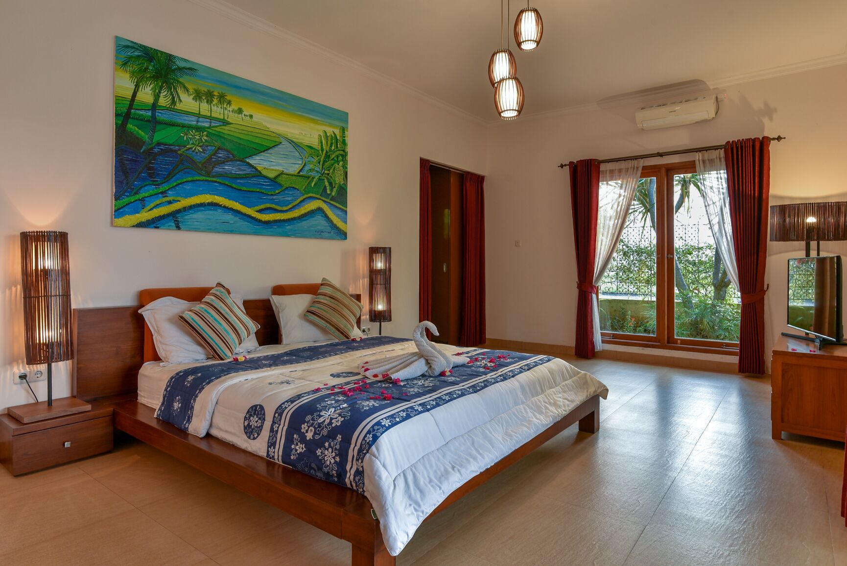 Un dormitorio amplio con una cama grande, un cuadro de un paisaje tropical, un televisor y un balcón con cortinas.