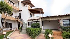 A multi-story building with a central staircase, a covered patio area, and a small garden with a palm tree and potted plants. - Irilena Apartments (Hersonissos)