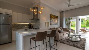 A modern kitchen with a central island, bar stools, a dining area, and a living space with a sofa and chairs. - Villa Shiva (Sukasada)