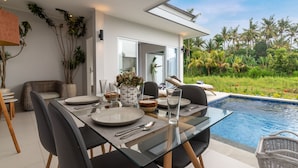 A modern dining area with a glass table, black chairs, and a view of a swimming pool and tropical garden. - Villa Shiva (Sukasada)