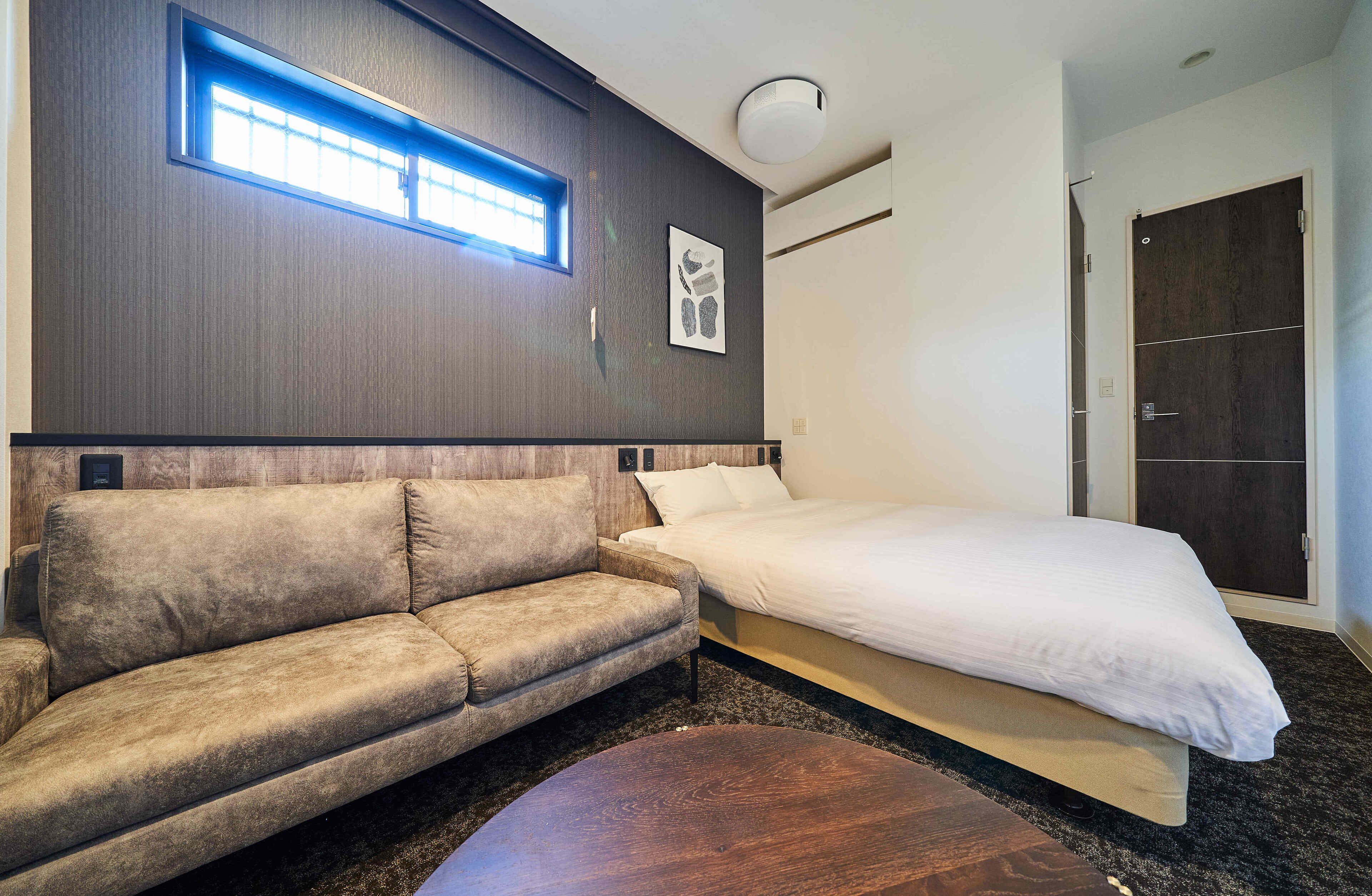 A modern hotel room with a sofa, a bed, a round coffee table, a window with blinds, and a door.