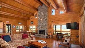 L’intérieur d’une cabane en rondins, avec une cheminée en pierre, des meubles en bois et de grandes fenêtres.