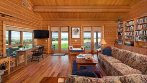L’intérieur d’une cabane en rondins, avec un plafond en bois, de grandes fenêtres, un canapé, une table basse, une bibliothèque et un téléviseur.