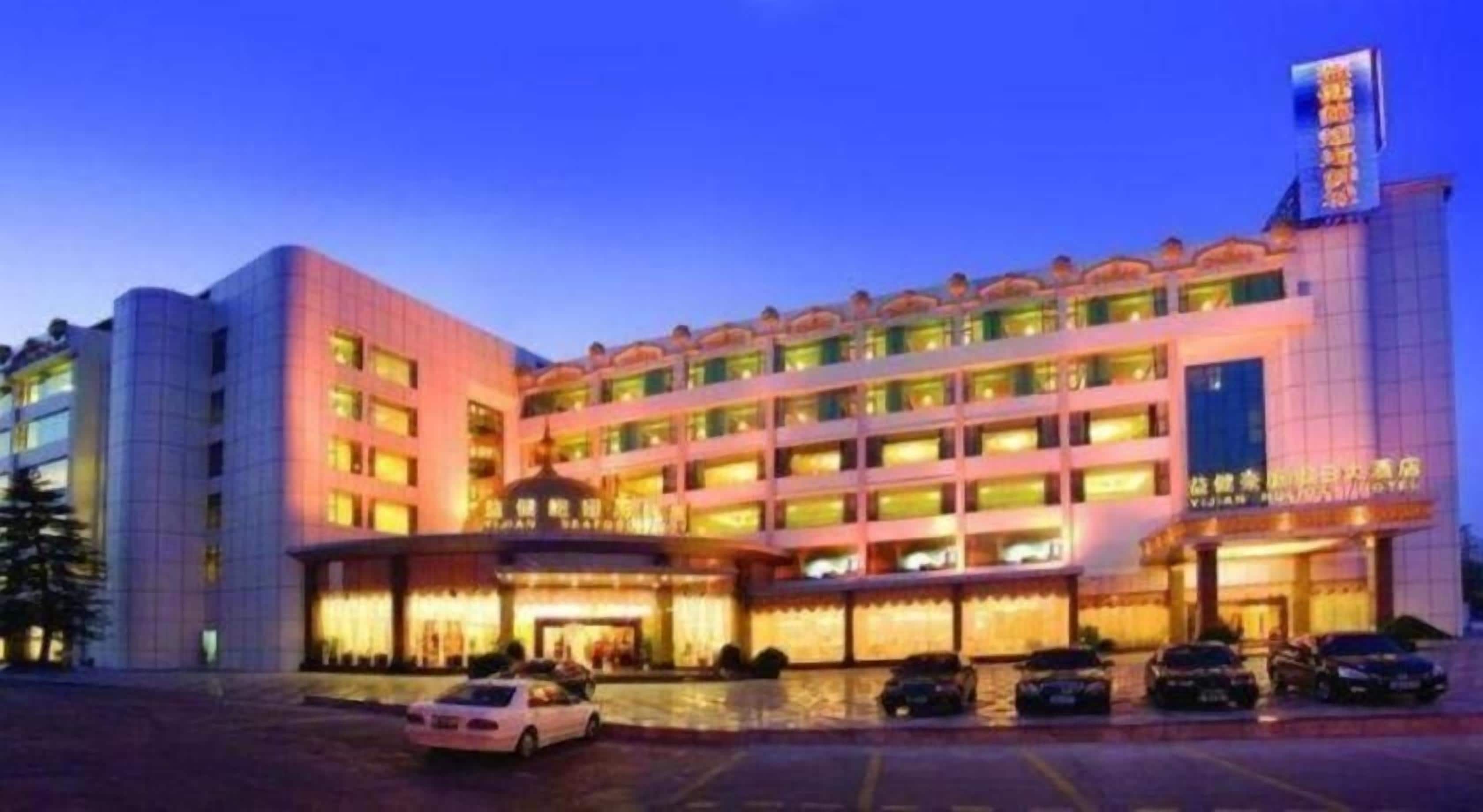 A multi-story hotel with a prominent entrance, multiple balconies, and a parking area with several cars.