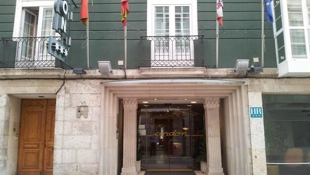 A hotel entrance with a glass door, a balcony with railings, and flags above. - Cordon (Burgos)