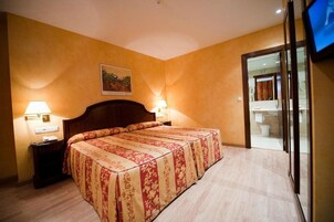 A hotel room with a large bed, two bedside lamps, a painting on the wall, and a view of the bathroom through a glass door.