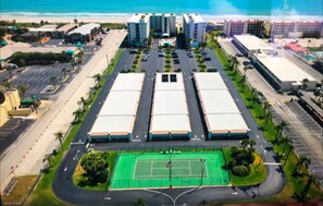 Vista aérea de un complejo hotelero con varias edificaciones, una cancha de tenis y zonas de estacionamiento.