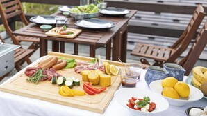 Una mesa de madera al aire libre con diversos alimentos, entre ellos carnes, verduras y frutas, además de un bol de ensalada.