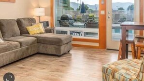 A living room with a sofa, a table, and a chair. There is a glass door leading to an outdoor area with a pool and umbrellas. - Chelan Cabana Chalet (chelan)