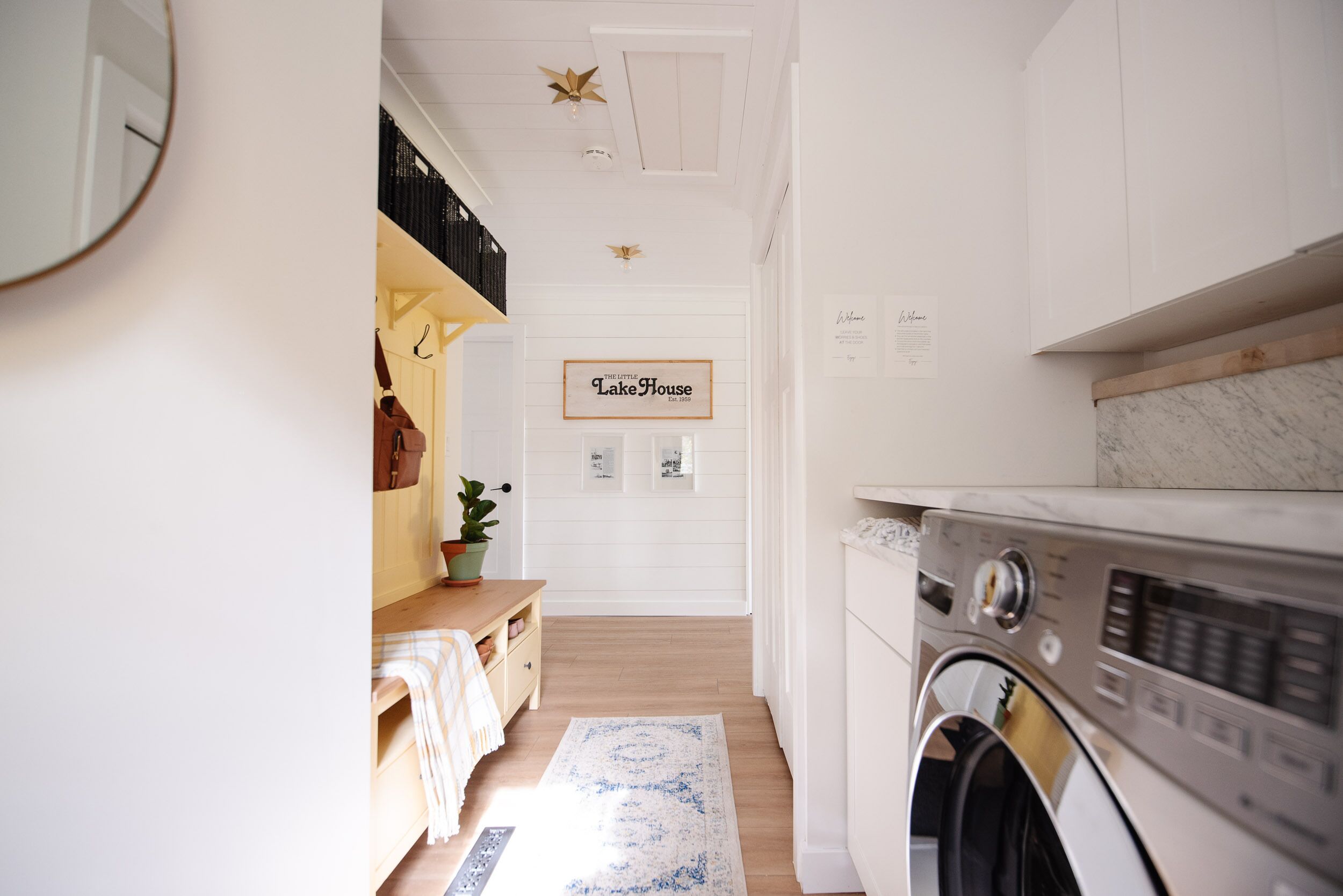 A laundry room with a washing machine, a wooden bench, a potted plant, and a sign that reads 'The Little Lake House.'