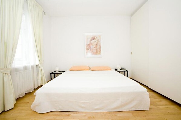 A bedroom with a bed, two nightstands, a framed picture on the wall, and sheer curtains.