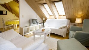 Een knus zolderkamertje met een bed, een slaapbank, een klein tafeltje met een bloem en een tv op een witte standaard.
