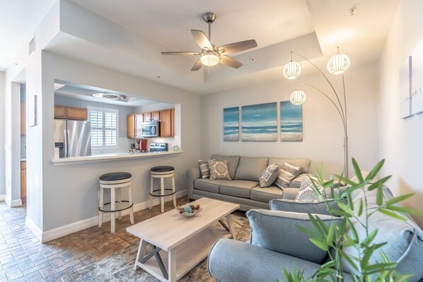 A modern living room with a sofa, coffee table, and bar stools. A kitchen with appliances and a window is visible in the background. - Fantastic Water Views | Newly Updated 3 Bed Town Home | Pet friendly | Sleeps 8 (Ruskin)