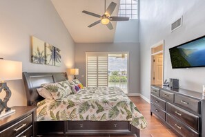 A bedroom with a bed, a ceiling fan, a TV, a window with blinds, and a view of the outdoors.