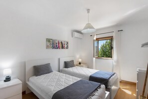 Un dormitorio con dos camas, una mesita de noche, una lámpara, equipo de aire acondicionado, una ventana con cortinas y un ventilador de techo.