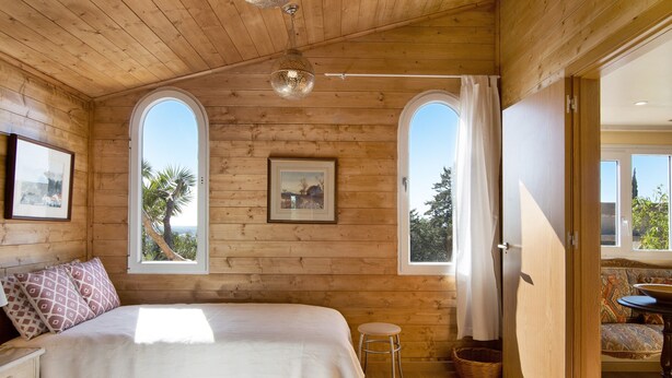 A wooden cabin bedroom with a bed, two windows, a picture on the wall, and a small stool. - Cabana Garden House with Shared Pool, Panoramic View & Wi-Fi (Loulé)