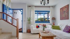 A living room with a staircase, a white sofa, a wooden coffee table, and a window with blue shutters. - Vacation Home 'Rosa' with Private Terrace, Private Garden and Wi-Fi (Yaiza)
