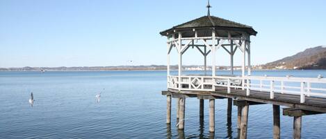 A gazebo on a pier extending into calm water, with a clear sky and distant land in the background.